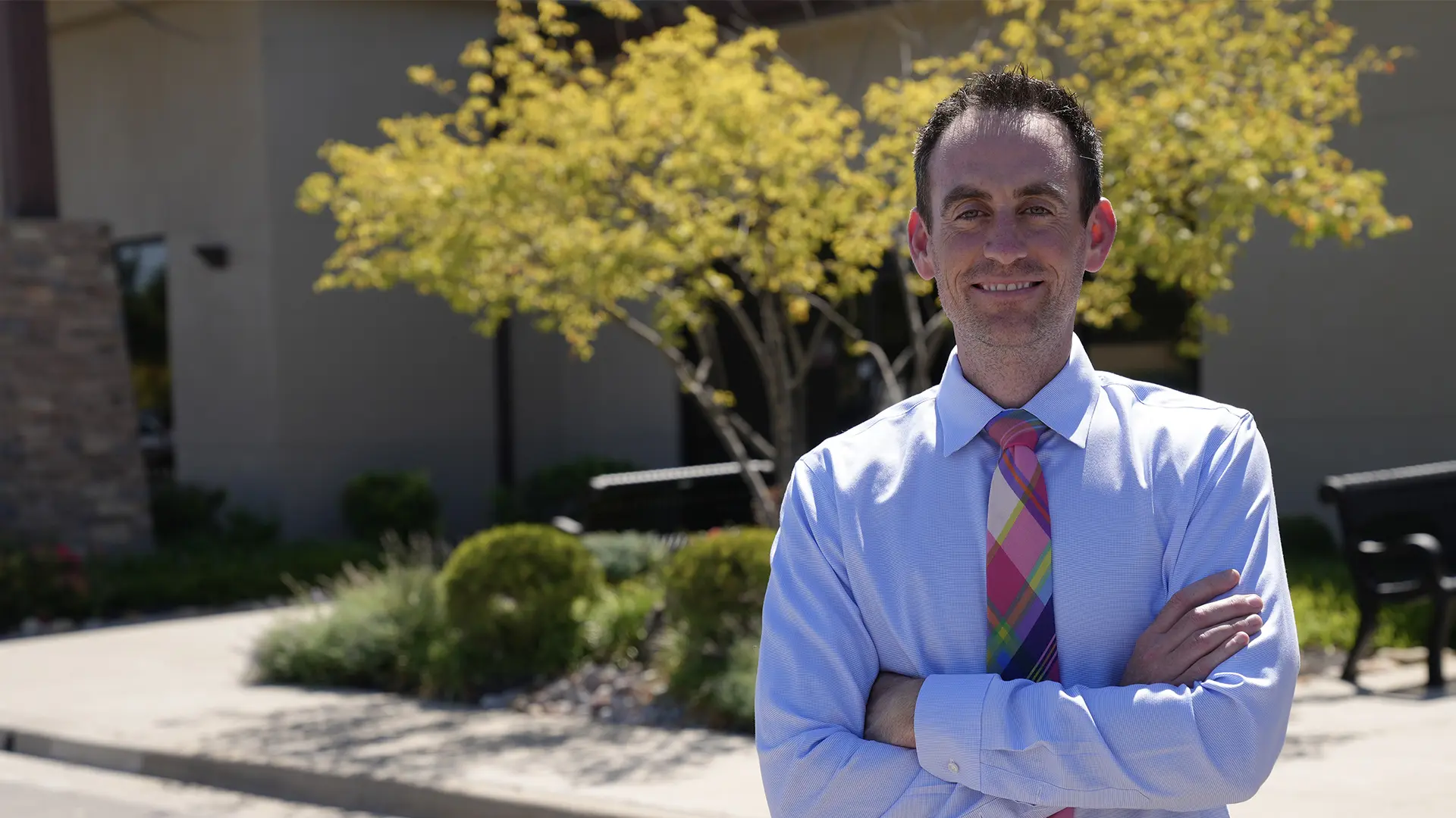 Smiling with arms crossed, Dr. Jeff Lenius stands outside Lenius Orthodontics in Overland Park, KS, by a building and tree.