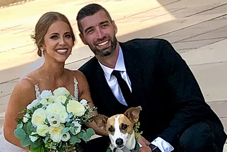 A bride and groom with a brown and white dog hold white flowers outside near Lenius Orthodontics in Overland Park, KS.