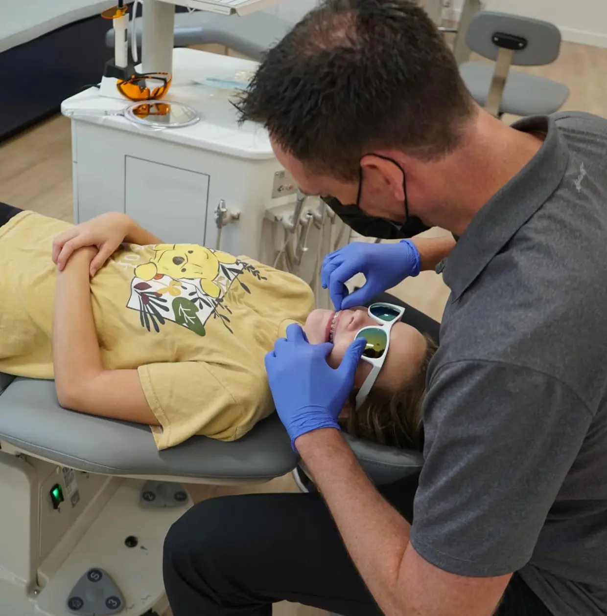 At Lenius Orthodontics in Overland Park, KS, a masked dental professional examines a reclining patient with sunglasses and representing personalized orthodontic excellence.