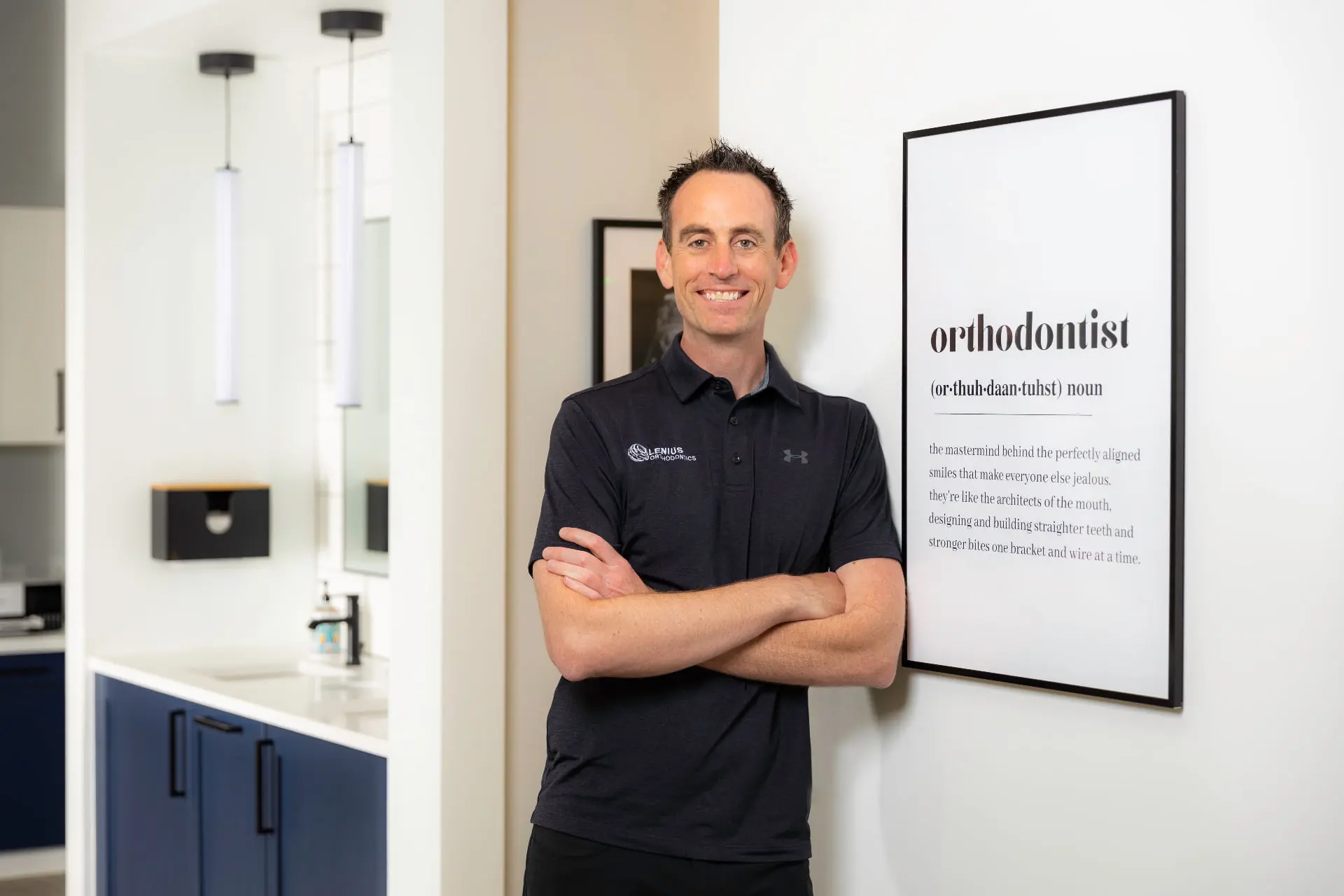 An orthodontics eexpert in a black polo shirt stands with arms crossed by a framed "orthodontist" sign at Lenius Orthodontics in Overland Park, KS.