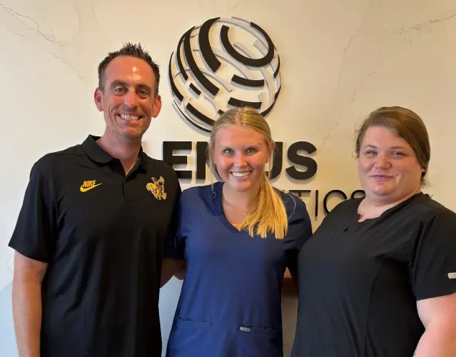 Three people smiling in front of a wall with a circular logo and partial text, represent for schedule Invisalign free consultation at Lenius Orthodontics in Overland Park, KS.