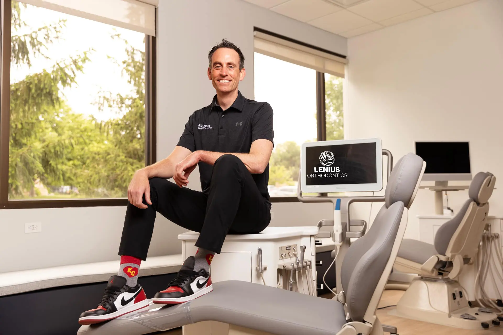 Dr. Jeff Lenius in a black shirt and sneakers sits on a dental chair, with "Lenius Orthodontics" in Overland Park, KS on a monitor.