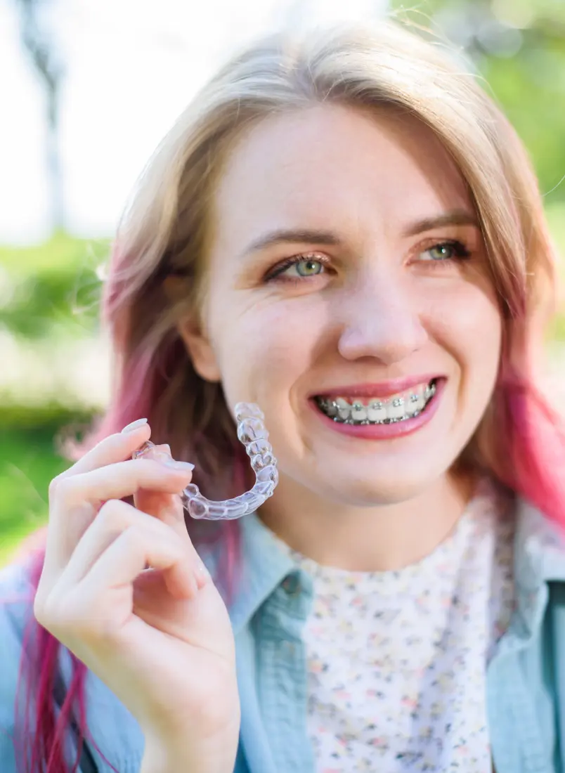 Smiling young woman with braces holds a braces outdoors on a sunny day at Lenius Orthodontics in Overland Park, KS.