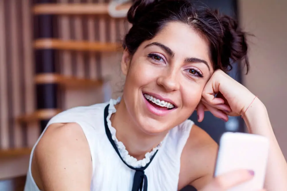 Smiling young woman with dark hair in buns and metal braces at Lenius Orthodontics in Overland Park, KS, holding a smartphone.