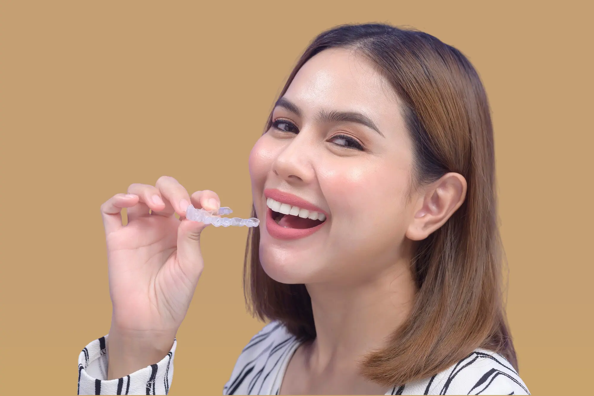 Smiling woman holds an affordable Invisalign aligner near her mouth, representing Lenius Orthodontics in Overland Park, KS.