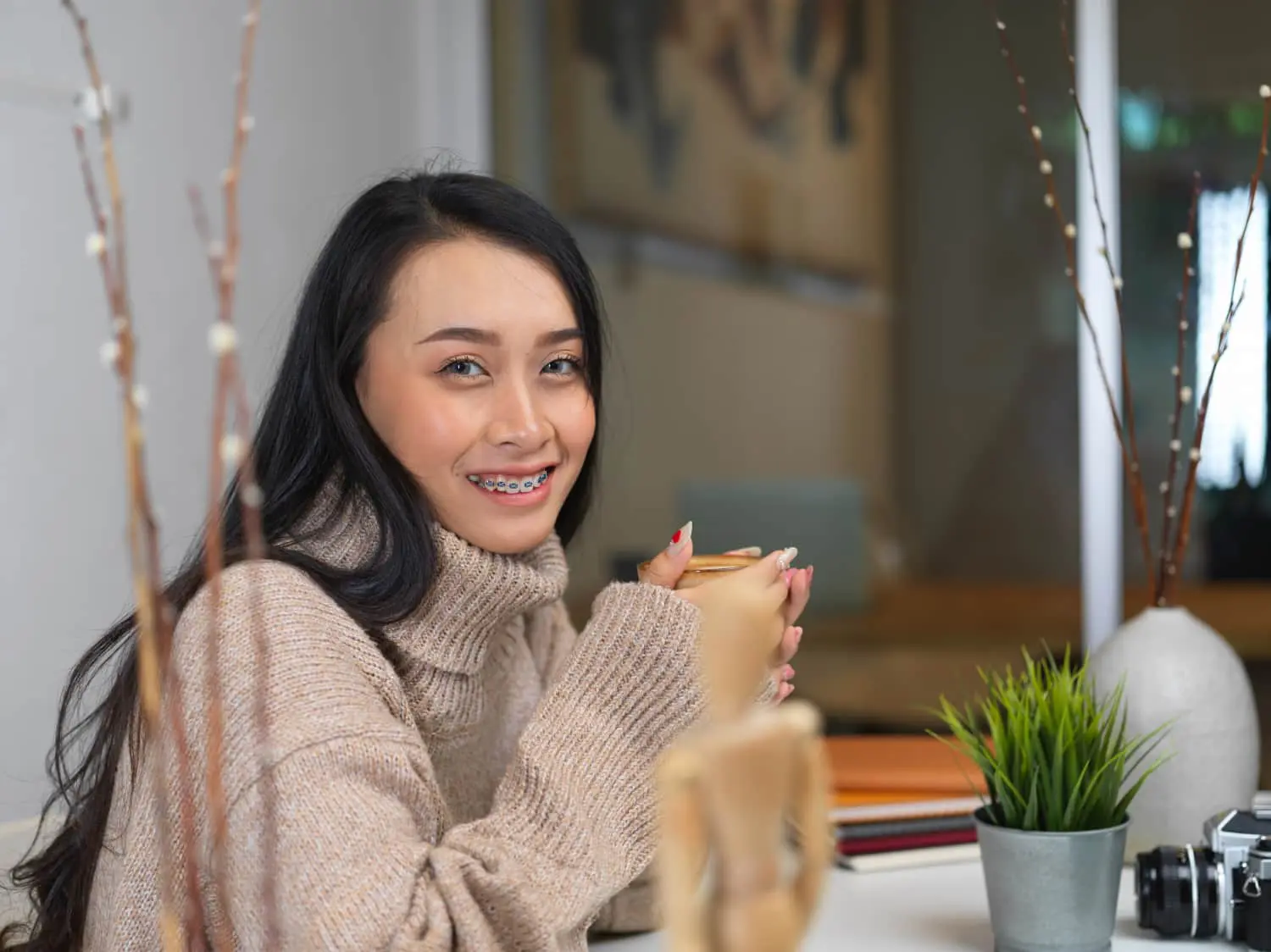 Smiling woman with braces at a desk, enjoying Lenius Orthodontics insurance benefits in Overland Park, KS; plants nearby.