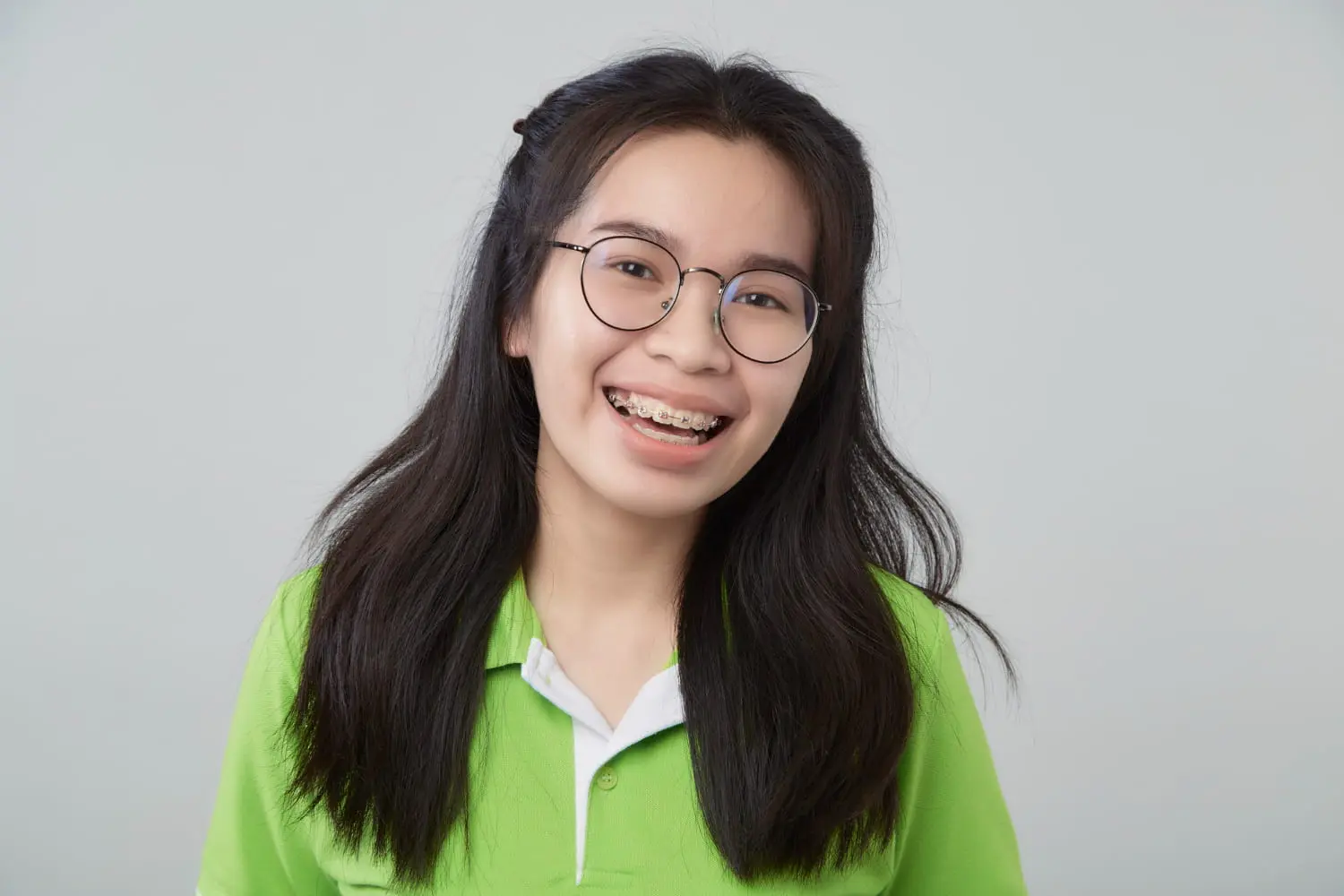 A young woman with long dark hair, glasses, and braces smiles confidently in a green shirt, benefiting from the Braces Payment Plan offered by Lenius Orthodontics in Overland Park, KS.