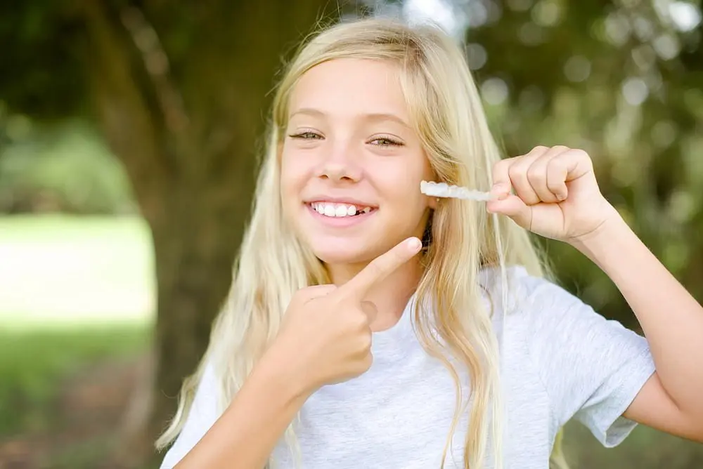 A smiling girl with long blonde hair points to clear aligners outdoors, from children orthodontist at Lenius Orthodontics in Overland Park, KS.