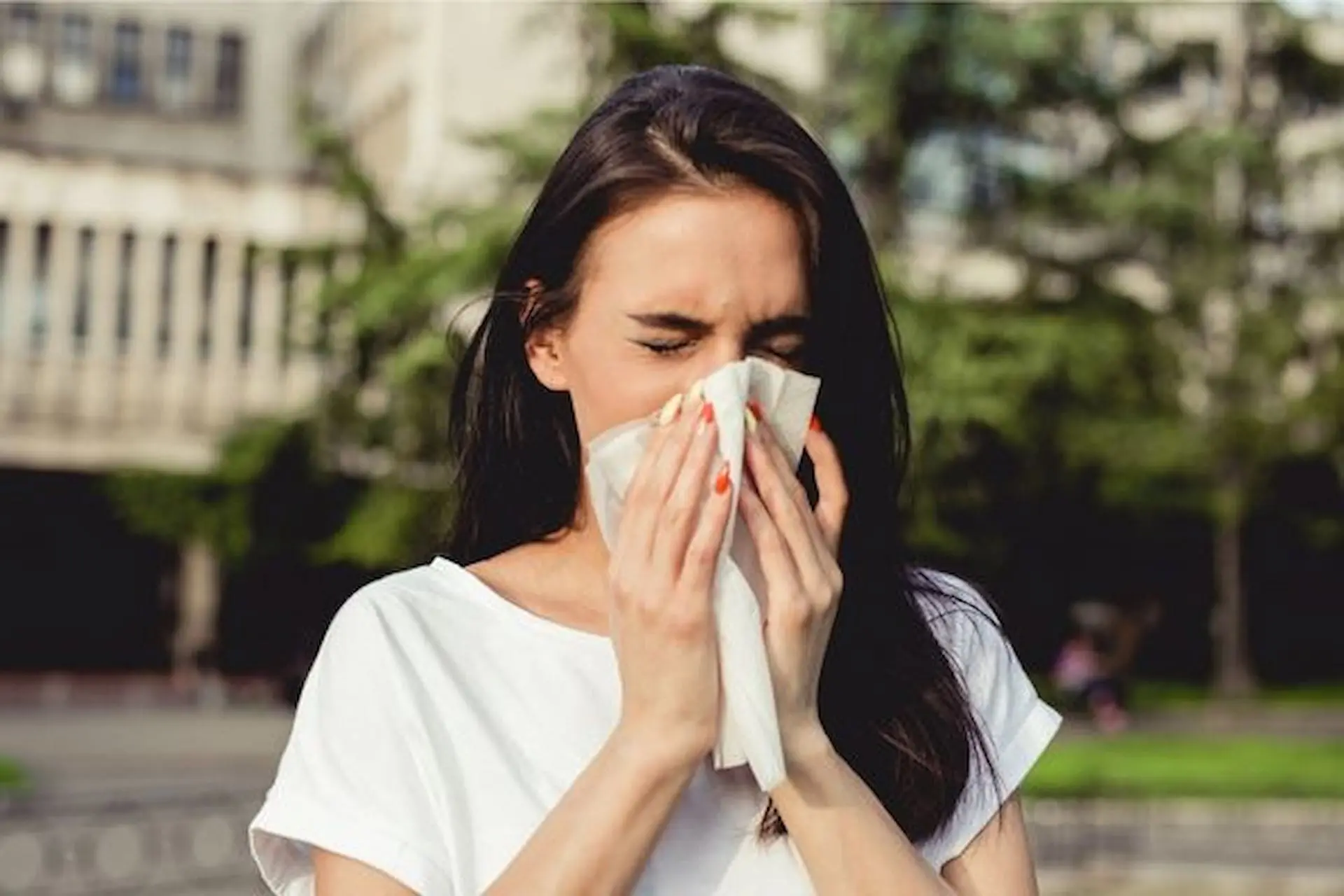 Sneezing into her hands, a woman in a white shirt stands outside, reminiscent of the dedicated mouth breather one might encounter at Lenius Orthodontics in Overland Park, KS.