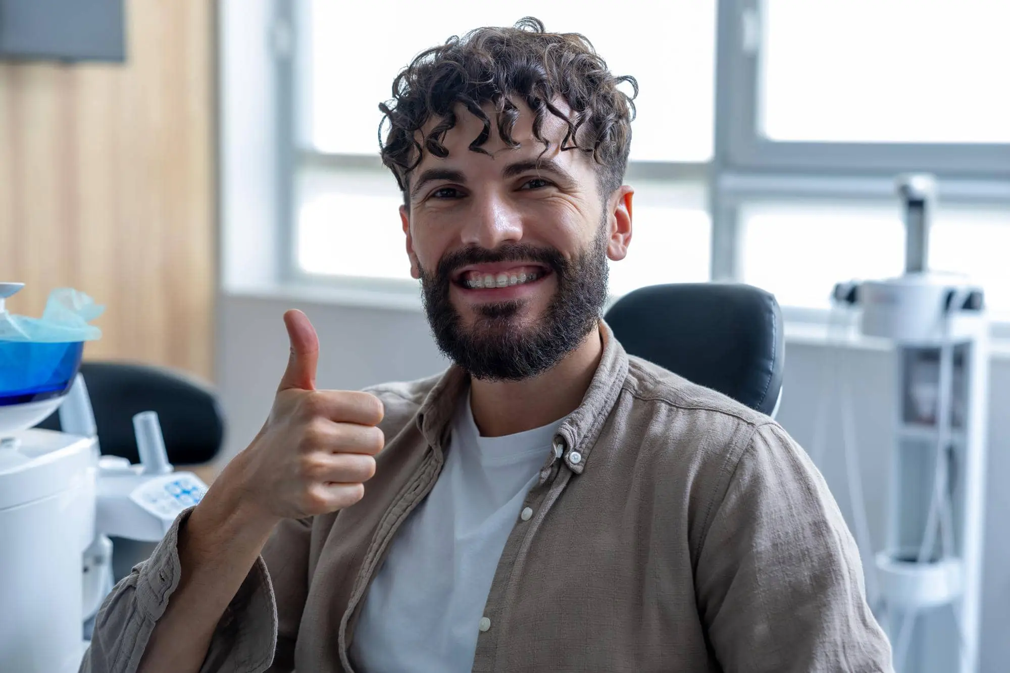 A smiling man with curly hair and a beard gives a thumbs-up near dental equipment at Lenius Orthodontics in Overland Park, KS, showcasing Types of Braces, Ceramic Braces options for patients.