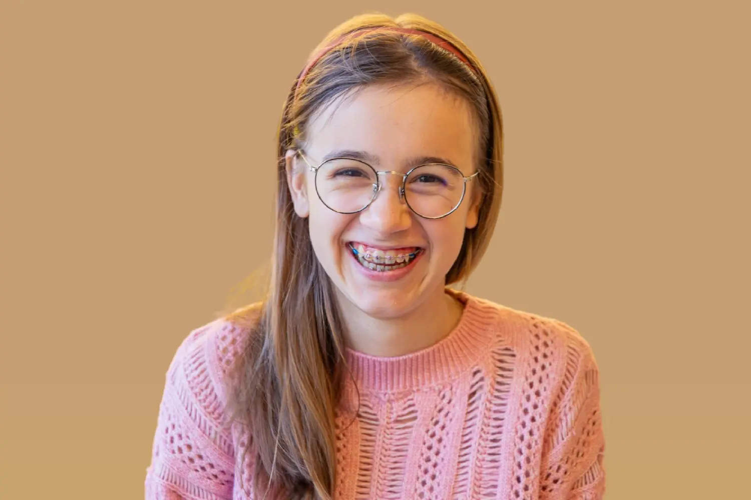 A smiling young person with braces and glasses wears a pink sweater, represent top-rated Children’s Orthodontists at Lenius Orthodontics in Overland Park, KS.