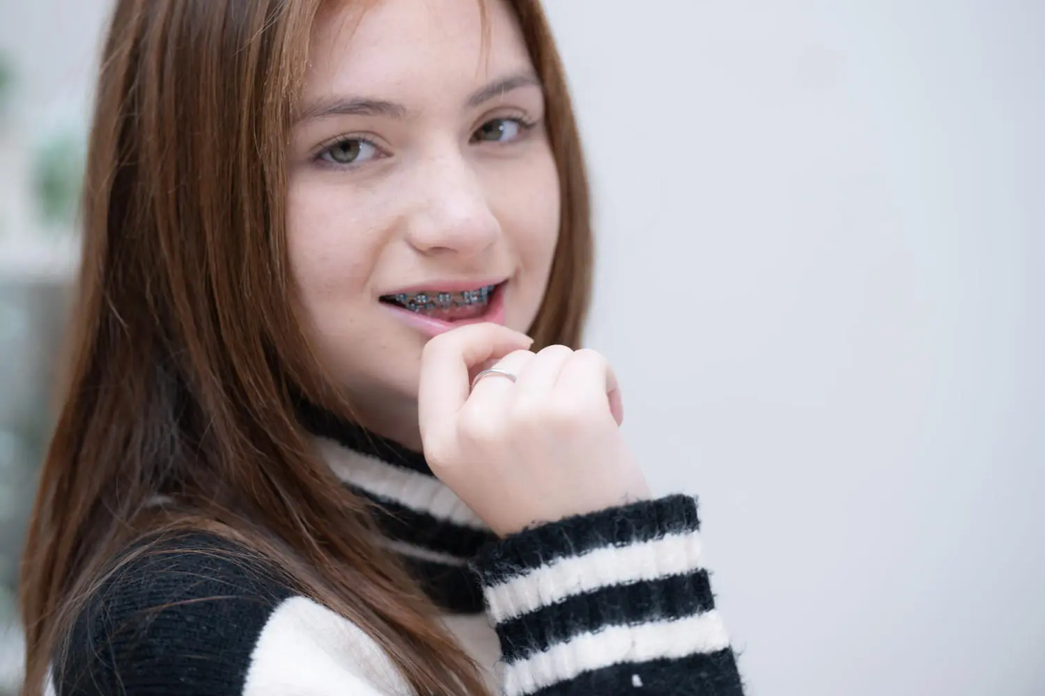 Smiling teen with long brown hair and metal braces, wearing a striped sweater, at Lenius Orthodontics in Overland Park, KS.