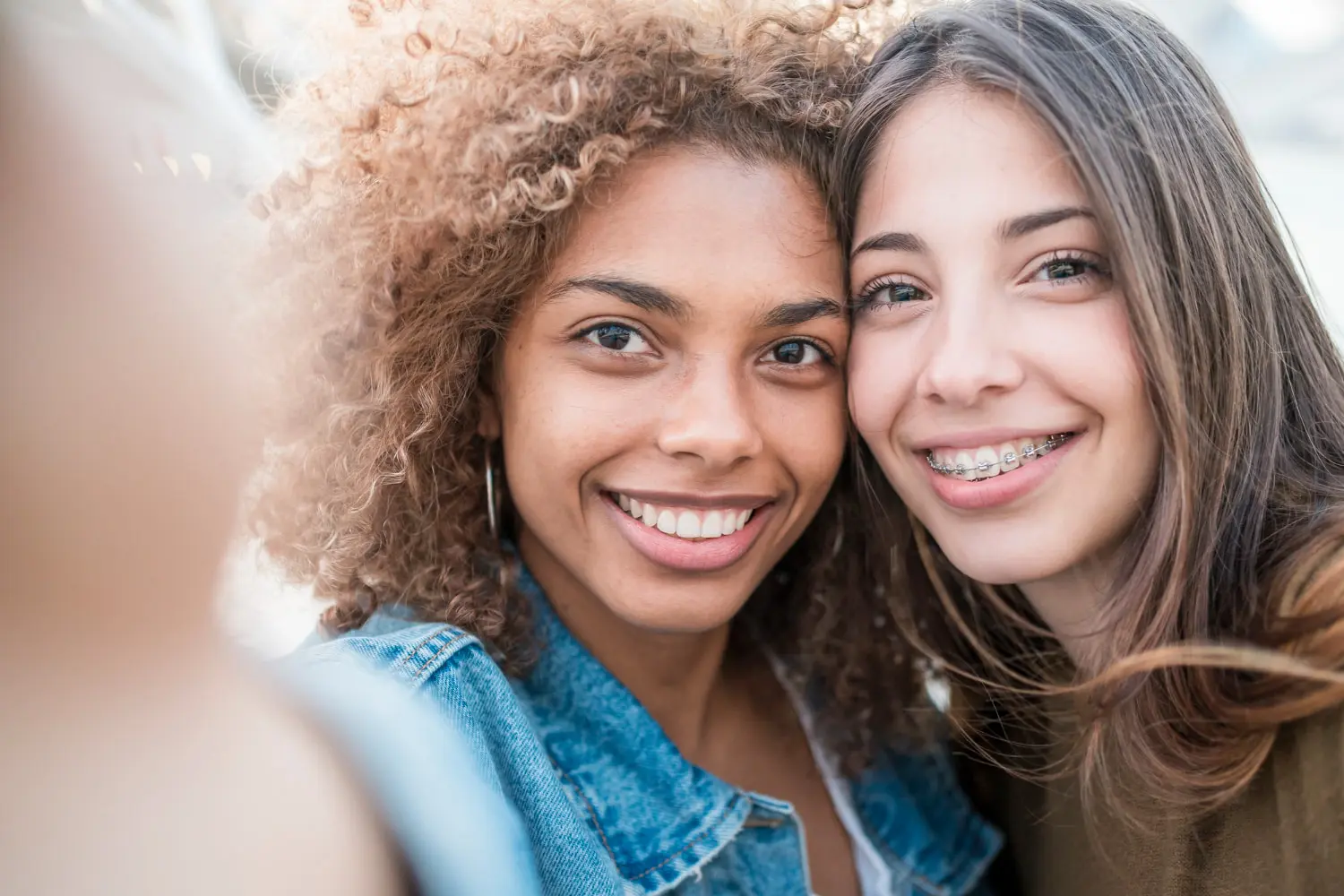 Two young women smile for a selfie at Lenius Orthodontics in Overland Park, KS—one with curly hair, one with affordable braces and straight hair.