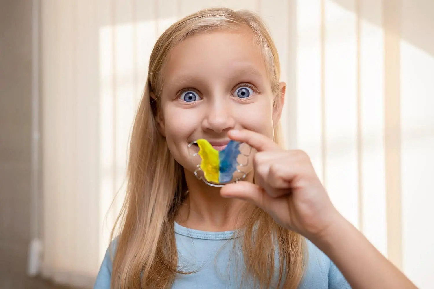 Smiling, a young girl with long blonde hair shows off colorful retainers from children orthodontist at Lenius Orthodontics in Overland Park, KS.