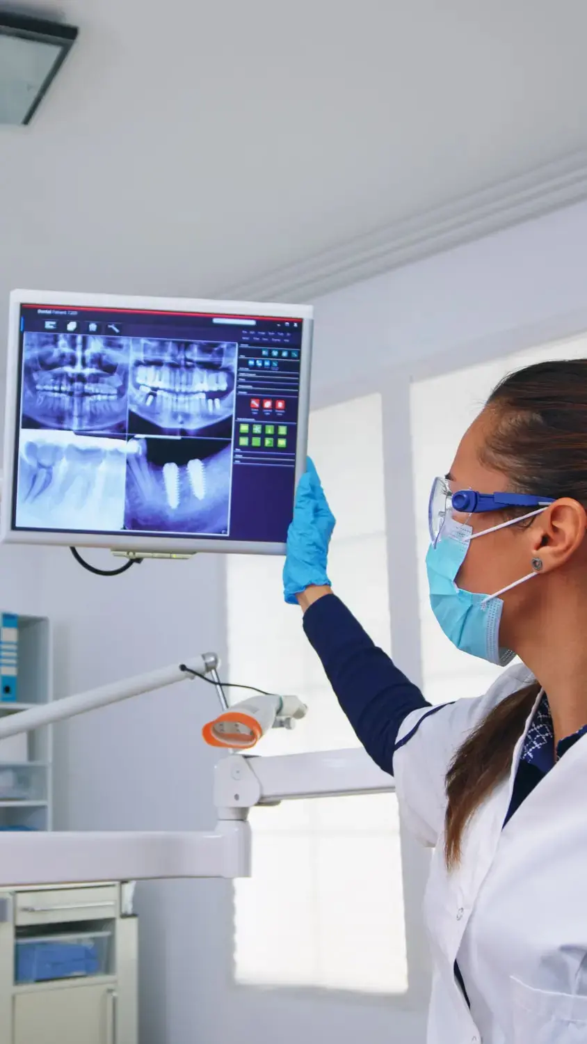 At Lenius Orthodontics in Overland Park, KS, a dentist in protective gear reviews dental X-rays on a clinic monitor explain about jaw surgery to patient.