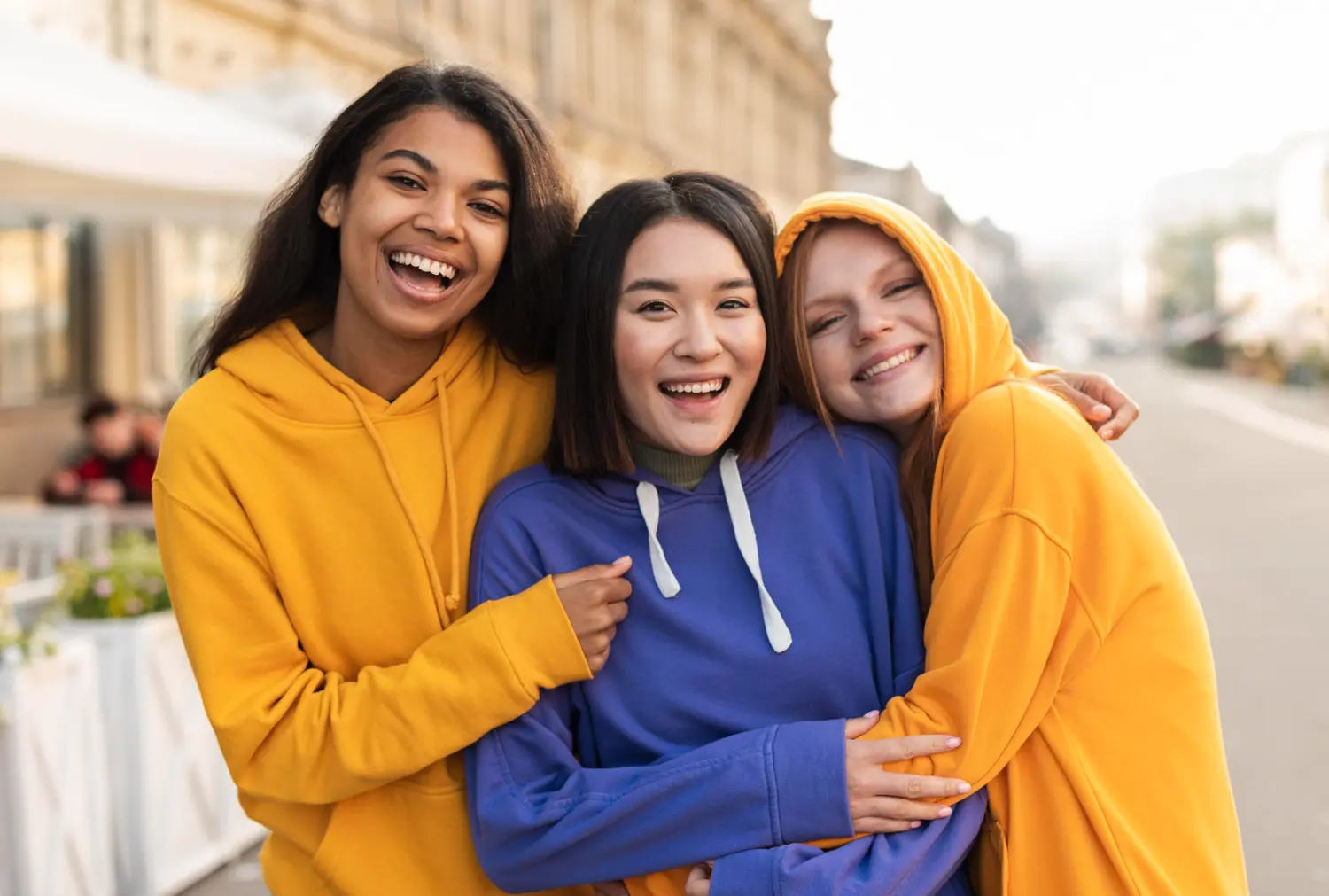 Smiling young women in colorful hoodies embrace on a city street, representing result affordable braces at Lenius Orthodontics in Overland Park, KS.