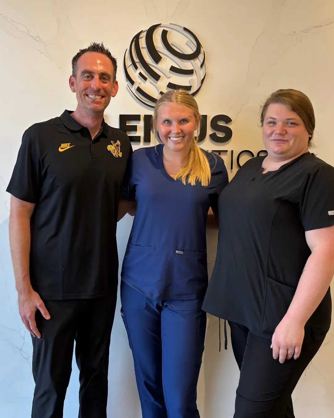 Two women in scrubs and Dr. Jeffrey Lenius is a orthodontic expert smiling in front of a Lenius Orthodontics logo wall in Overland Park, KS.