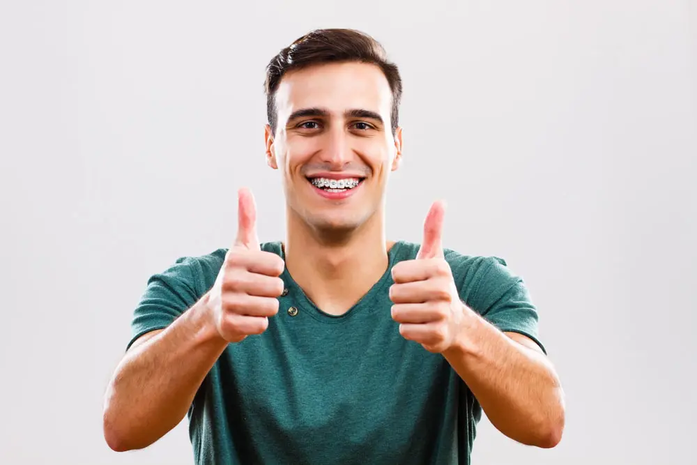 A young man in a green shirt with braces smiles and gives two thumbs up, highlighting Adult Orthodontic Treatment at Lenius Orthodontics in Overland Park, KS.