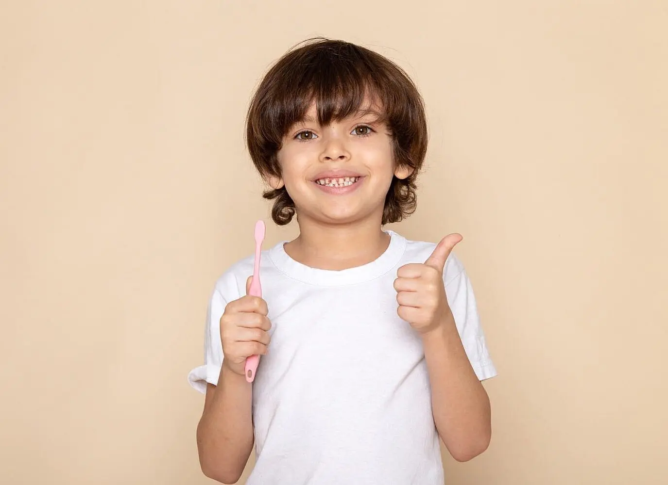 A smiling young boy in a white shirt represent overbite, underbite, crossbite and spacing Problems at Lenius Orthodontics in Overland Park, KS holds a pink toothbrush and gives a thumbs up.
