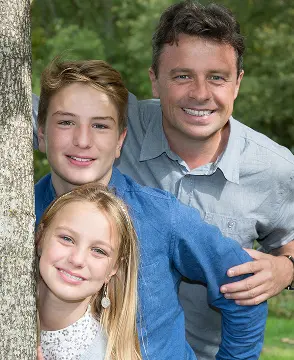Smiling together near a tree, a man show overbite with a teenage boy and young girl pose outside at Lenius Orthodontics in Overland Park, KS.