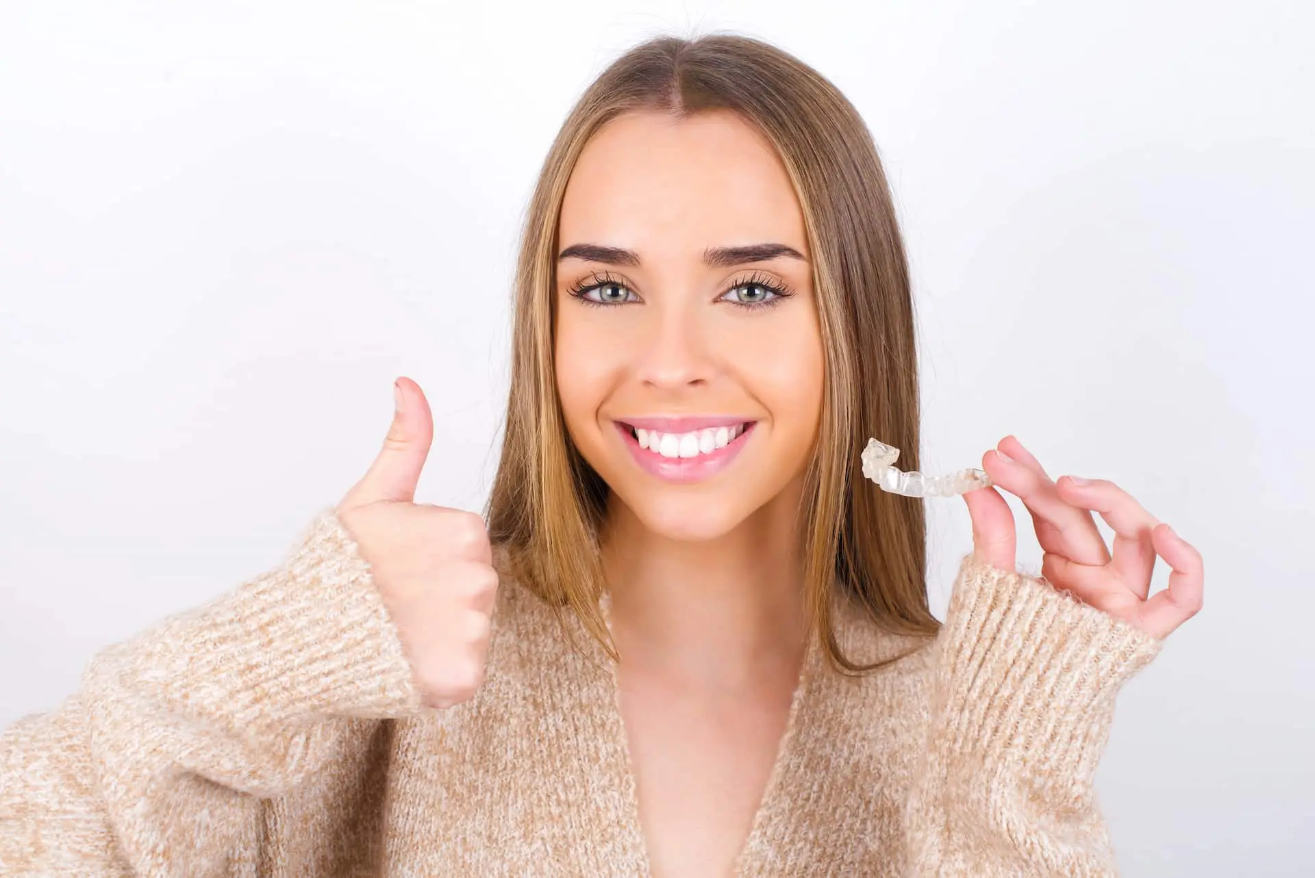 Smiling young woman shows Affordable Invisalign aligner and thumbs up, promoting Lenius Orthodontics in Overland Park, KS, on white background.