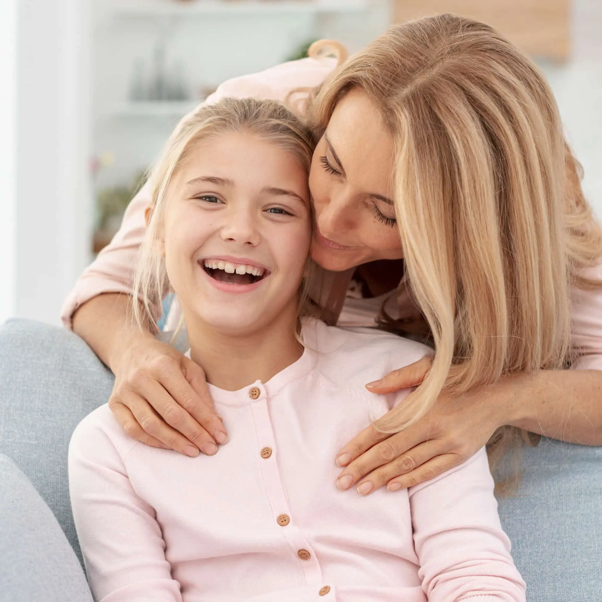 A woman hugs a smiling girl on a bright couch, reflecting Lenius Orthodontics' affordable Invisalign smiles in Overland Park, KS.