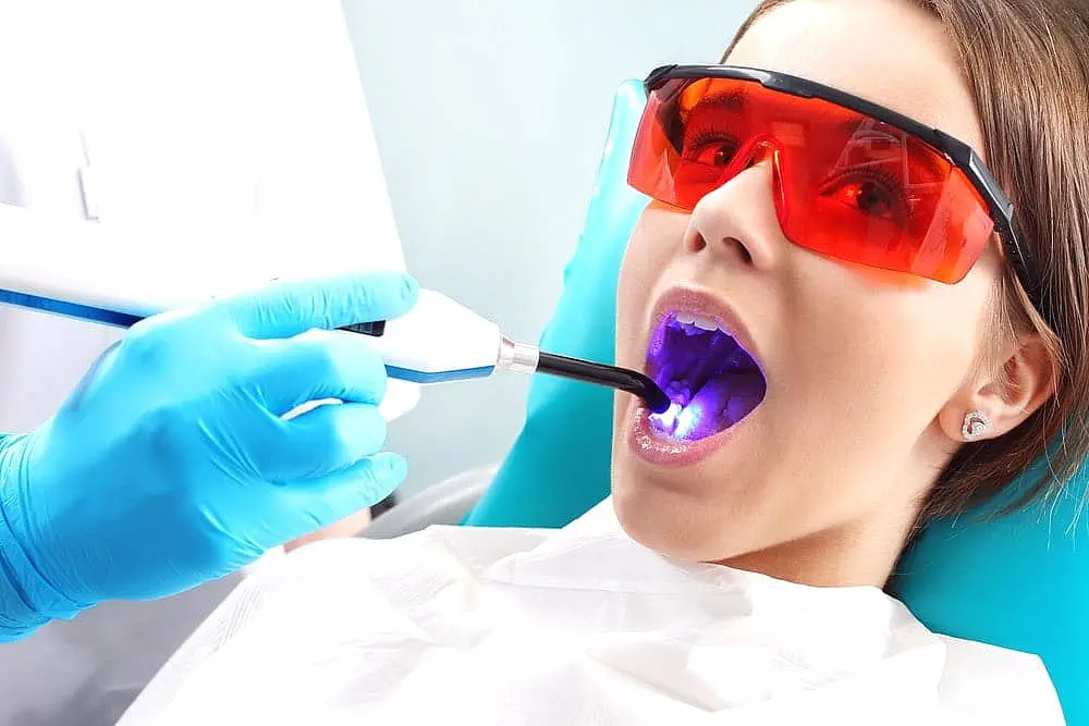 At Lenius Orthodontics in Overland Park, KS, a dentist uses a blue light on a patient wearing red glasses during Invisalign vs Braces care.
