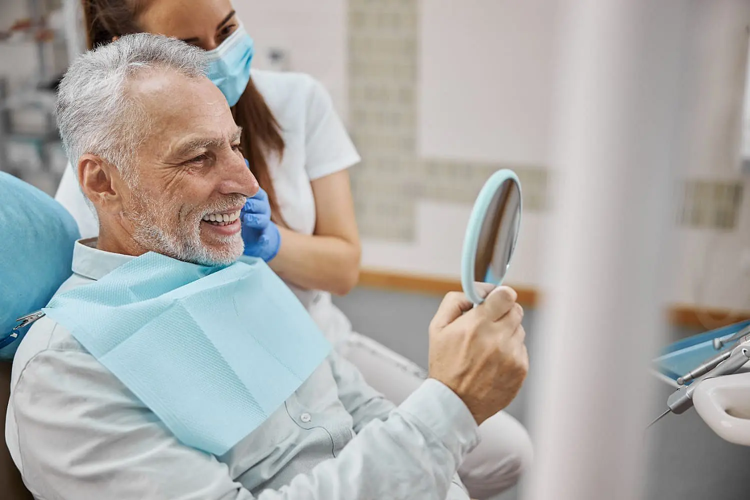 An older man smiles at his reflection in a handheld mirror show best Adult Orthodontics at Lenius Orthodontics in Overland Park, KS, with a masked dental professional nearby.