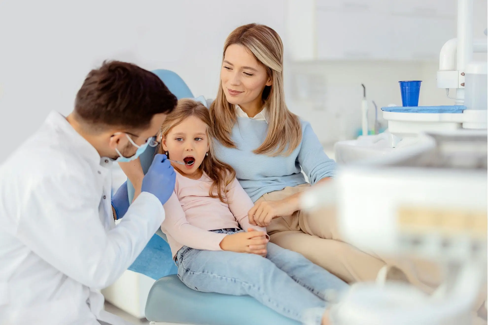 At Lenius Orthodontics in Overland Park, KS, a dentist checks a young girl's mouth as she sits on her mother's lap for represent long-term management and prognosis tongue thrust treatment.