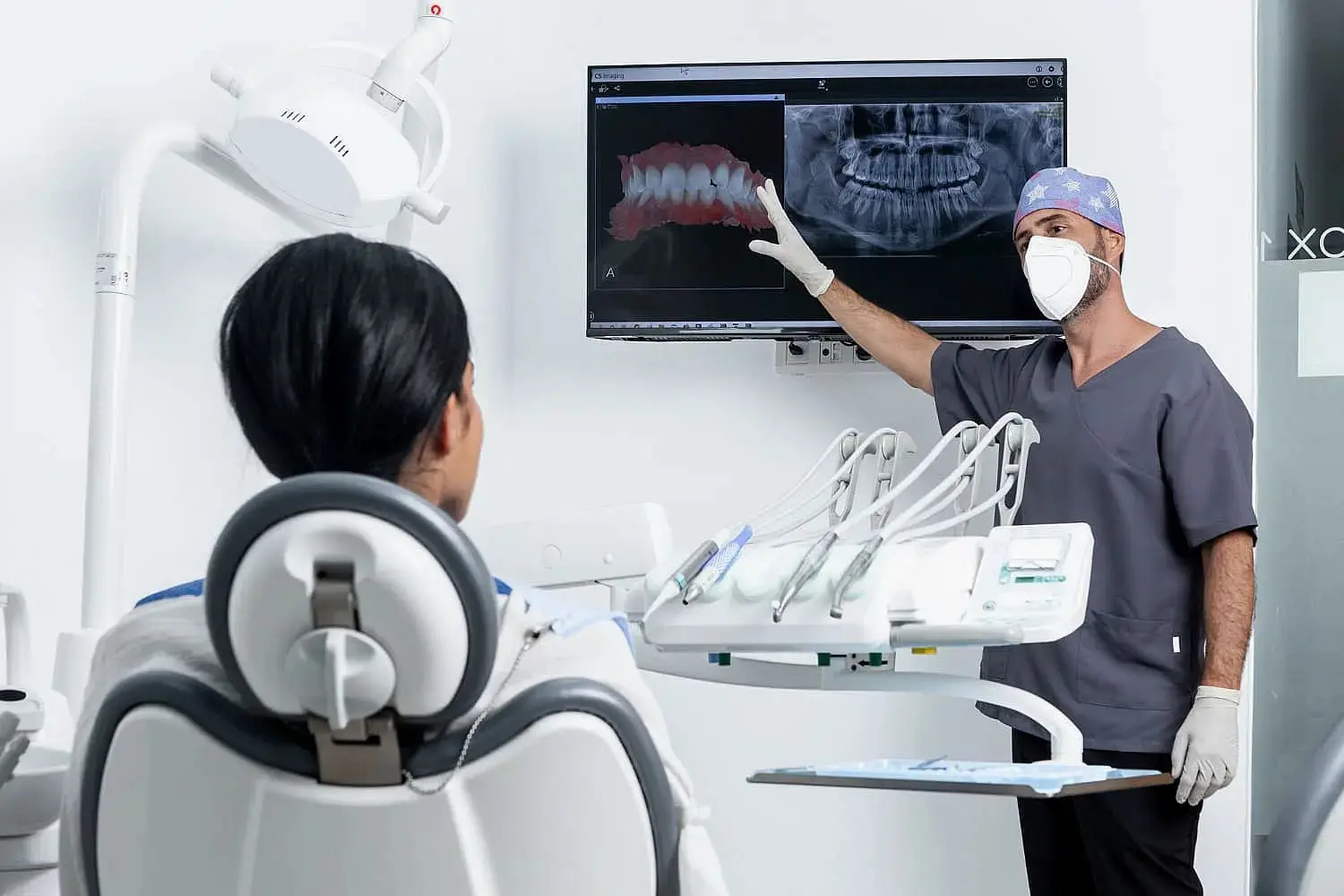 At Lenius Orthodontics in Overland Park, KS, a dentist in scrubs and a mask reviews dental X-rays on a monitor with a patient explain about benefits of jaw surgery.