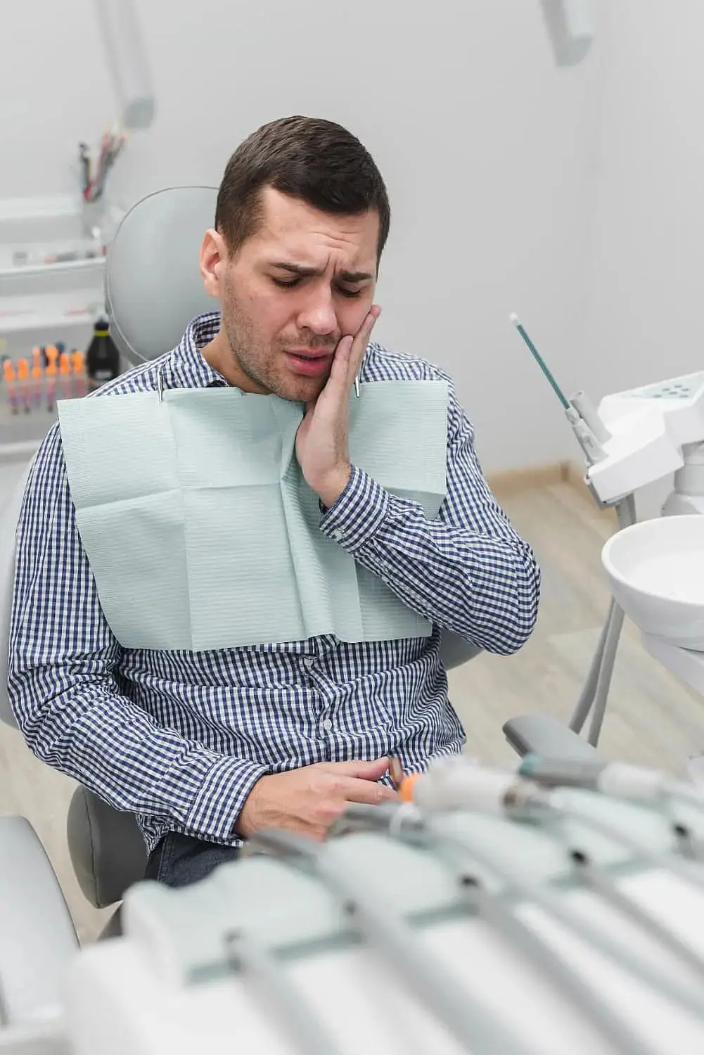 A man in a dental chair at Lenius Orthodontics in Overland Park, KS holds his cheek and winces, indicating dental discomfort need for Jaw Surgery.