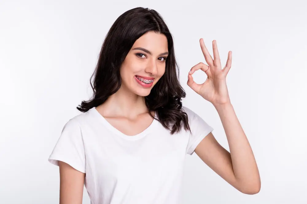 Smiling young woman with metal braces from Lenius Orthodontics in Overland Park, KS makes an "OK" gesture against a light background.