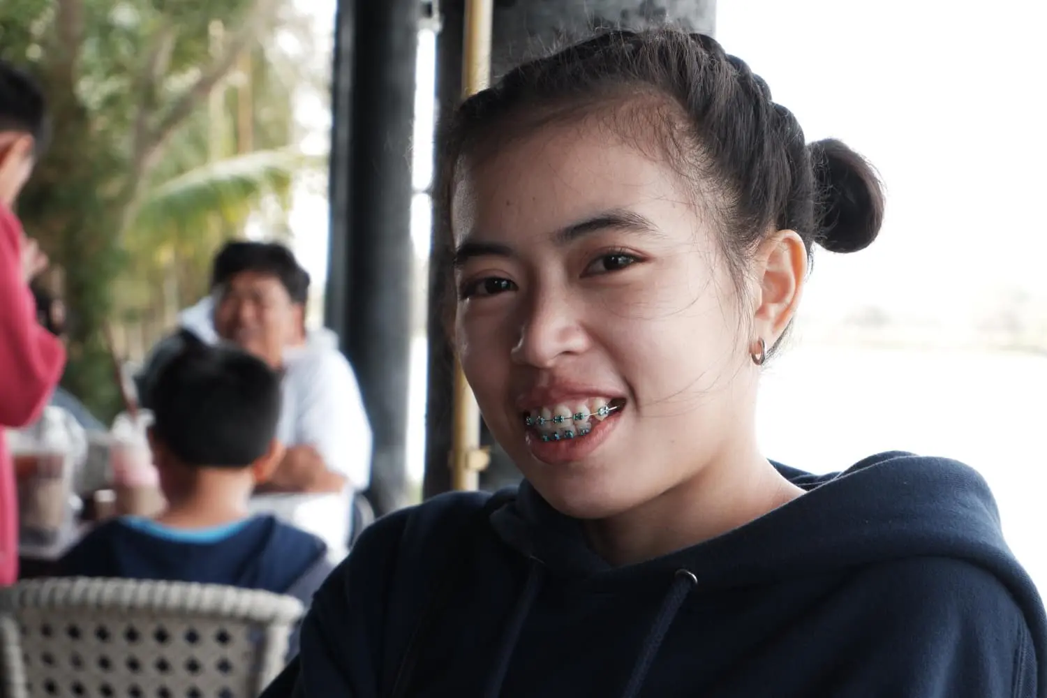 Smiling with metal braces, a young woman sits outdoors at Lenius Orthodontics in Overland Park, KS; people gather behind her.