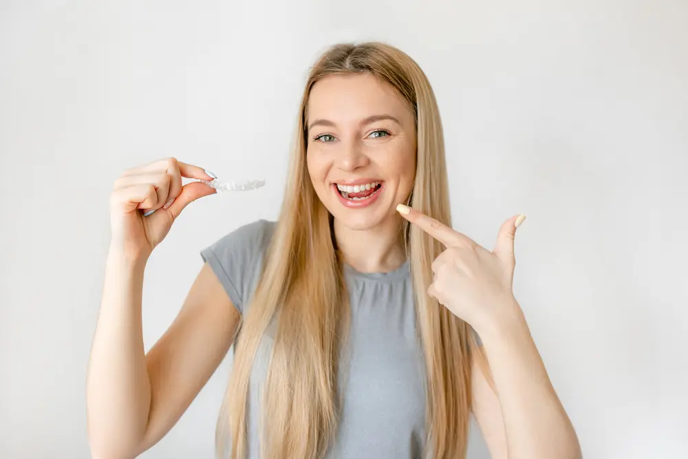 A smiling woman with long blonde hair points to her teeth and holds an Affordable Invisalign aligner at Lenius Orthodontics in Overland Park, KS.
