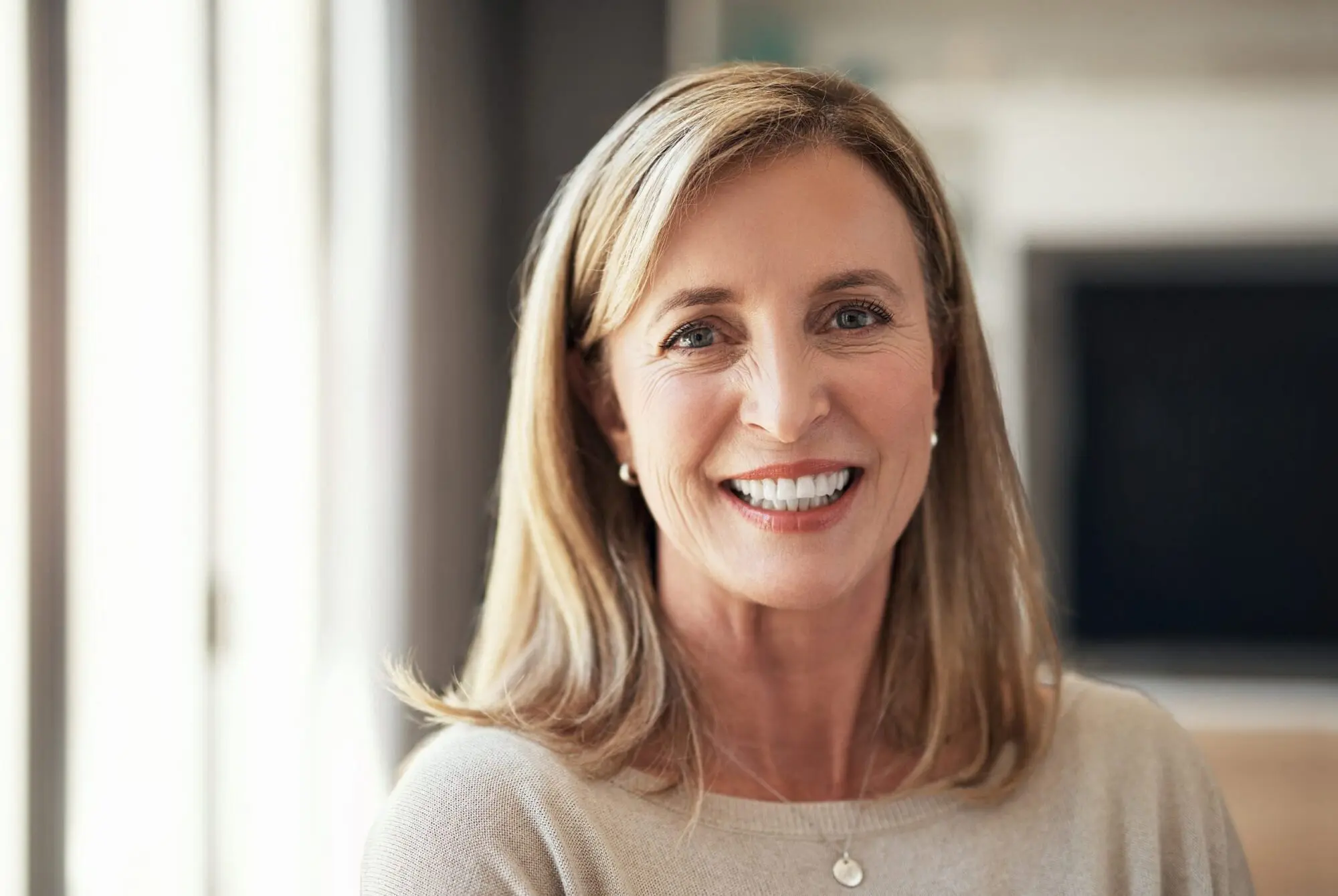 A woman with shoulder-length blonde hair smiles show result best affordable braces from Lenius Orthodontics in Overland Park, KS, wearing a light sweater and necklace.