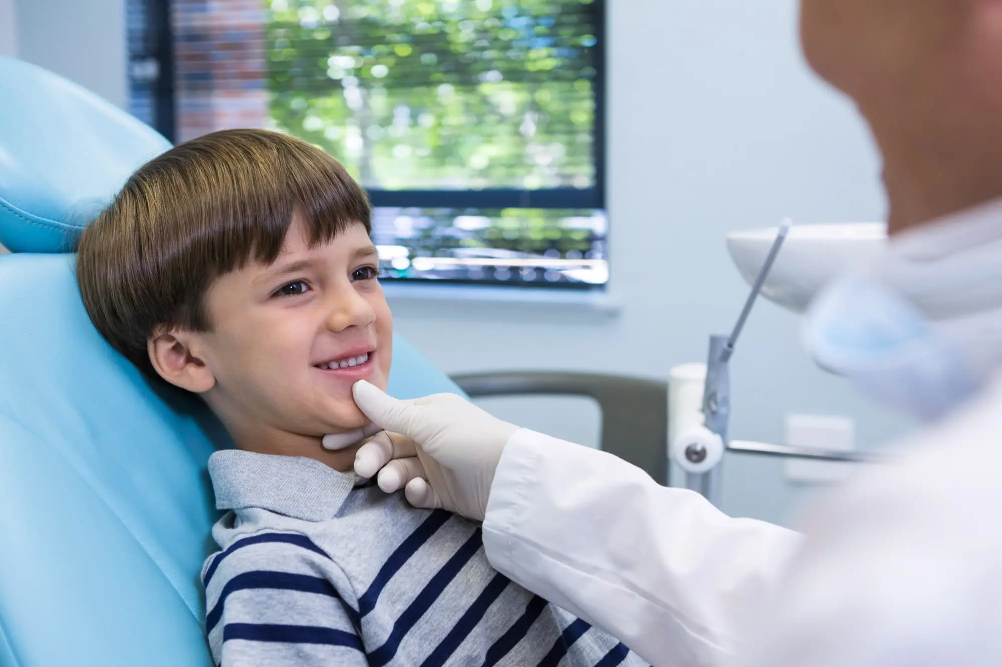 A young boy smiles in a dental chair as a gloved children orthodontist examines him show importance of early evaluation dental care at Lenius Orthodontics in Overland Park, KS.