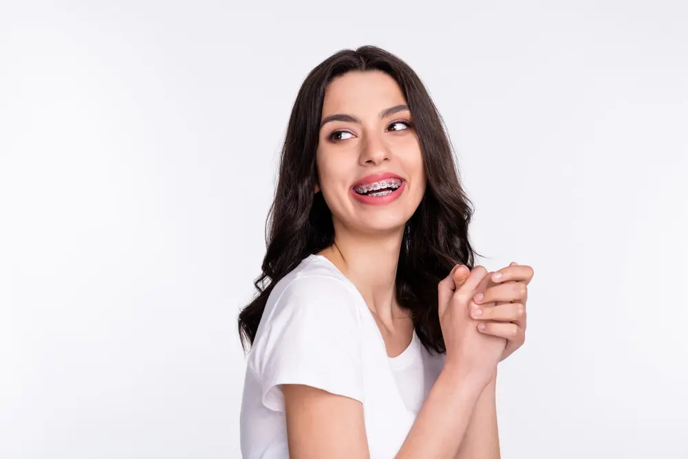Smiling woman with long brown hair and braces, in a white t-shirt, confident after learning about Braces Cost at Lenius Orthodontics in Overland Park, KS.