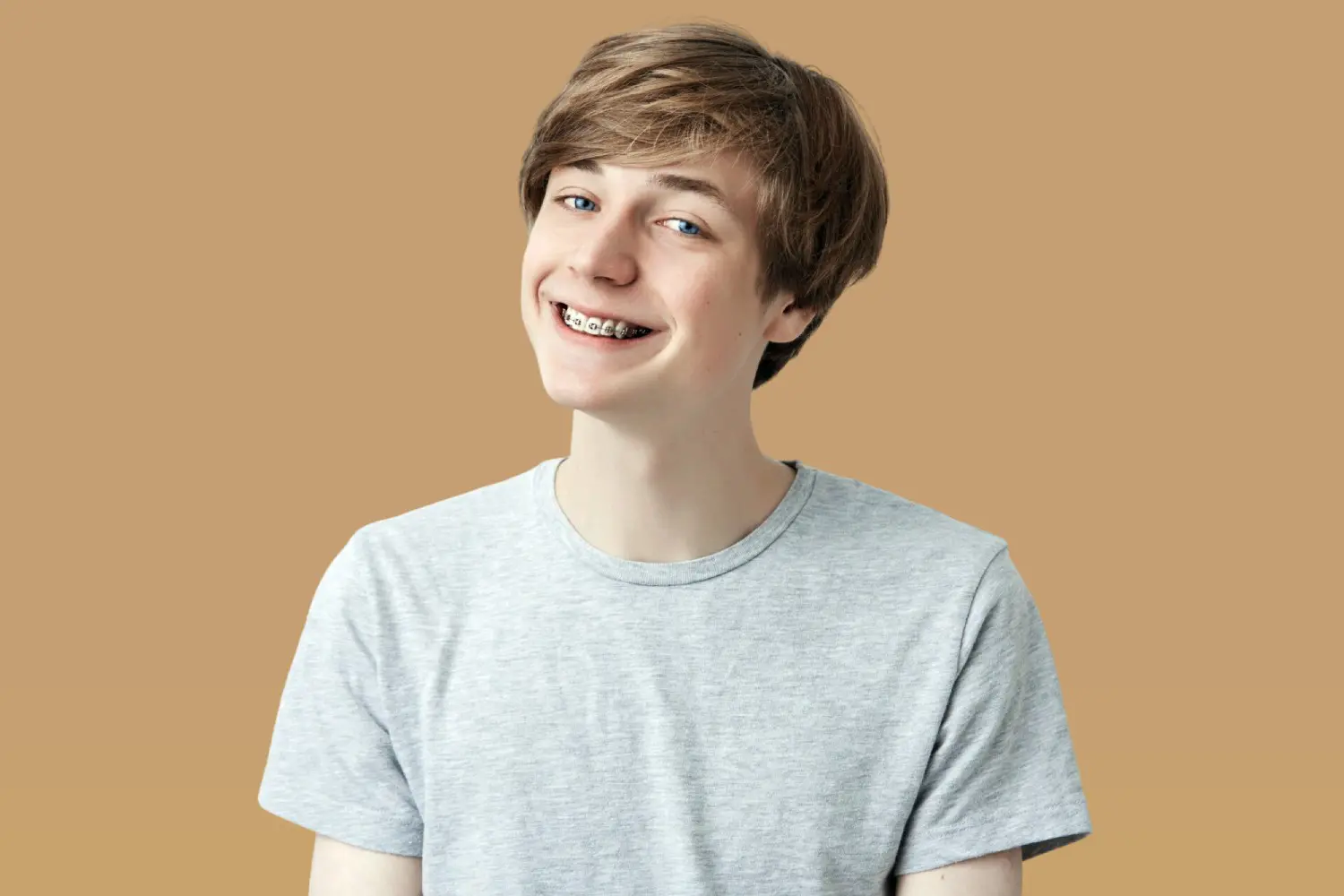 A teenage boy with braces smiles at the camera, showcasing different Types of Braces, for Lenius Orthodontics in Overland Park, KS, while wearing a gray t-shirt.