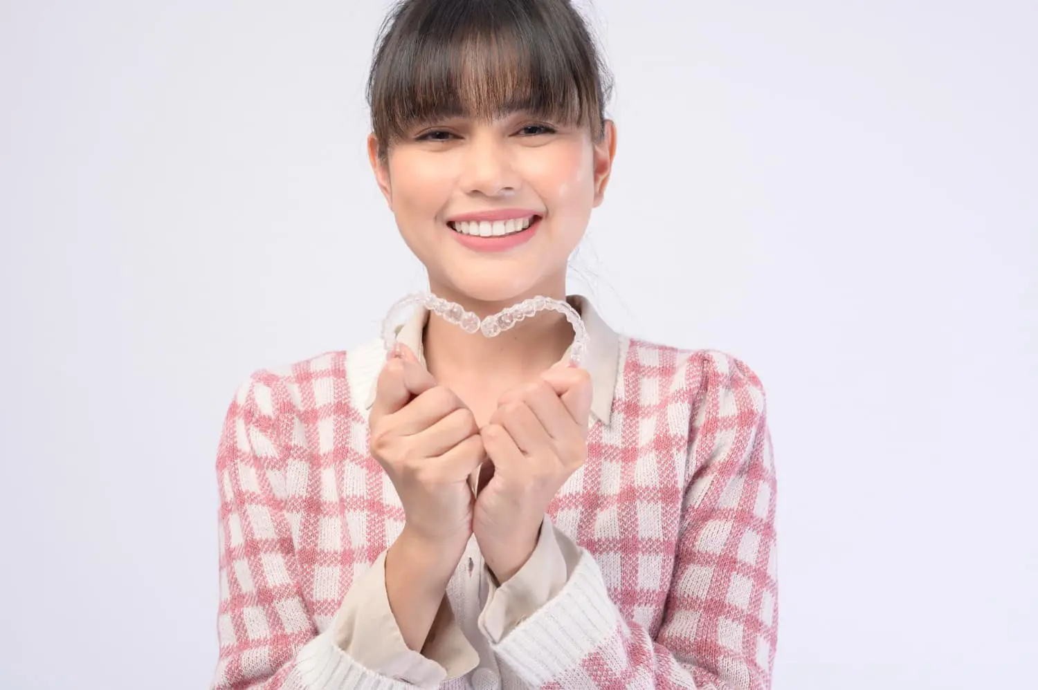 A smiling woman in a pink and white checkered sweater holds an aligner, highlighting Lenius Orthodontics Invisalign Payment Plan in Overland Park, KS.