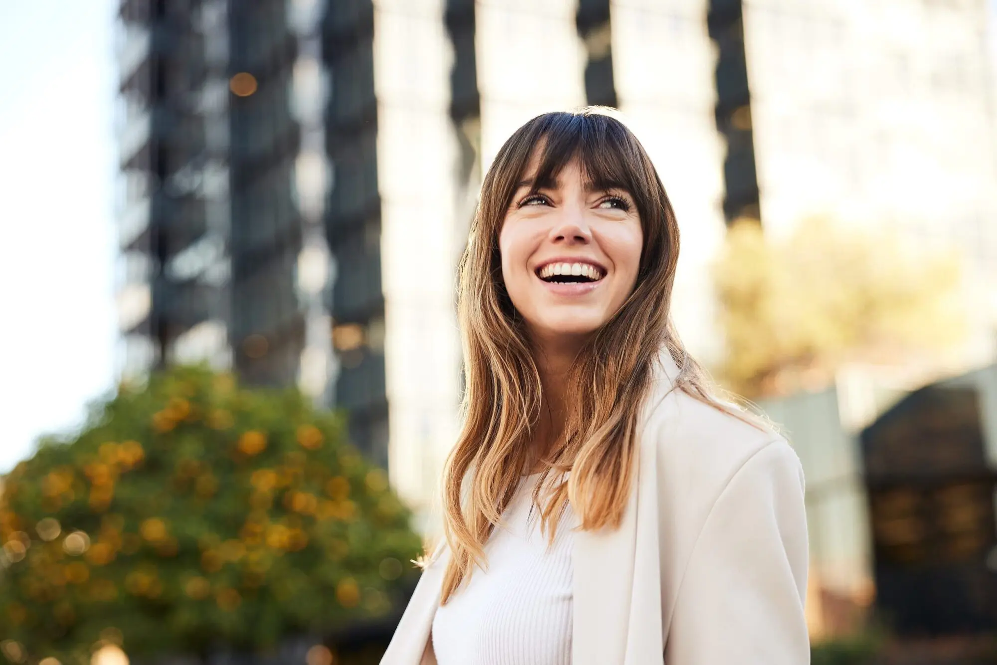 Smiling woman with long brown hair stands outside represent affordable braces from Lenius Orthodontics in Overland Park, KS, with greenery and sunlight behind her.