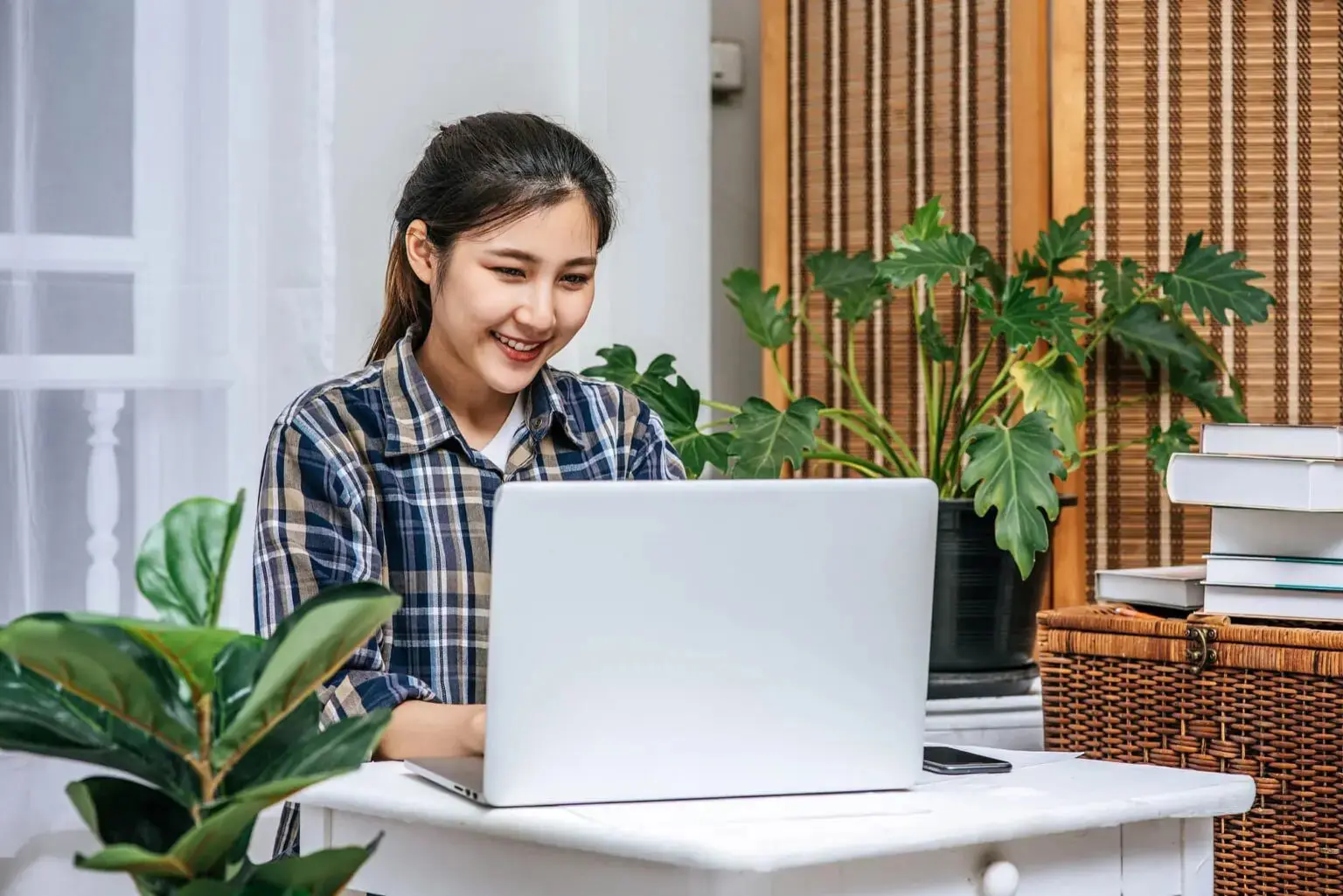 At home in Overland Park, KS, a woman works on her laptop by houseplants looking for affordable braces at Lenius Orthodontics.
