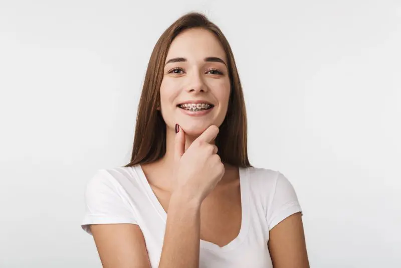 Smiling young woman in a white t-shirt and braces, hand on chin, representing affordable braces from Lenius Orthodontics in Overland Park, KS.