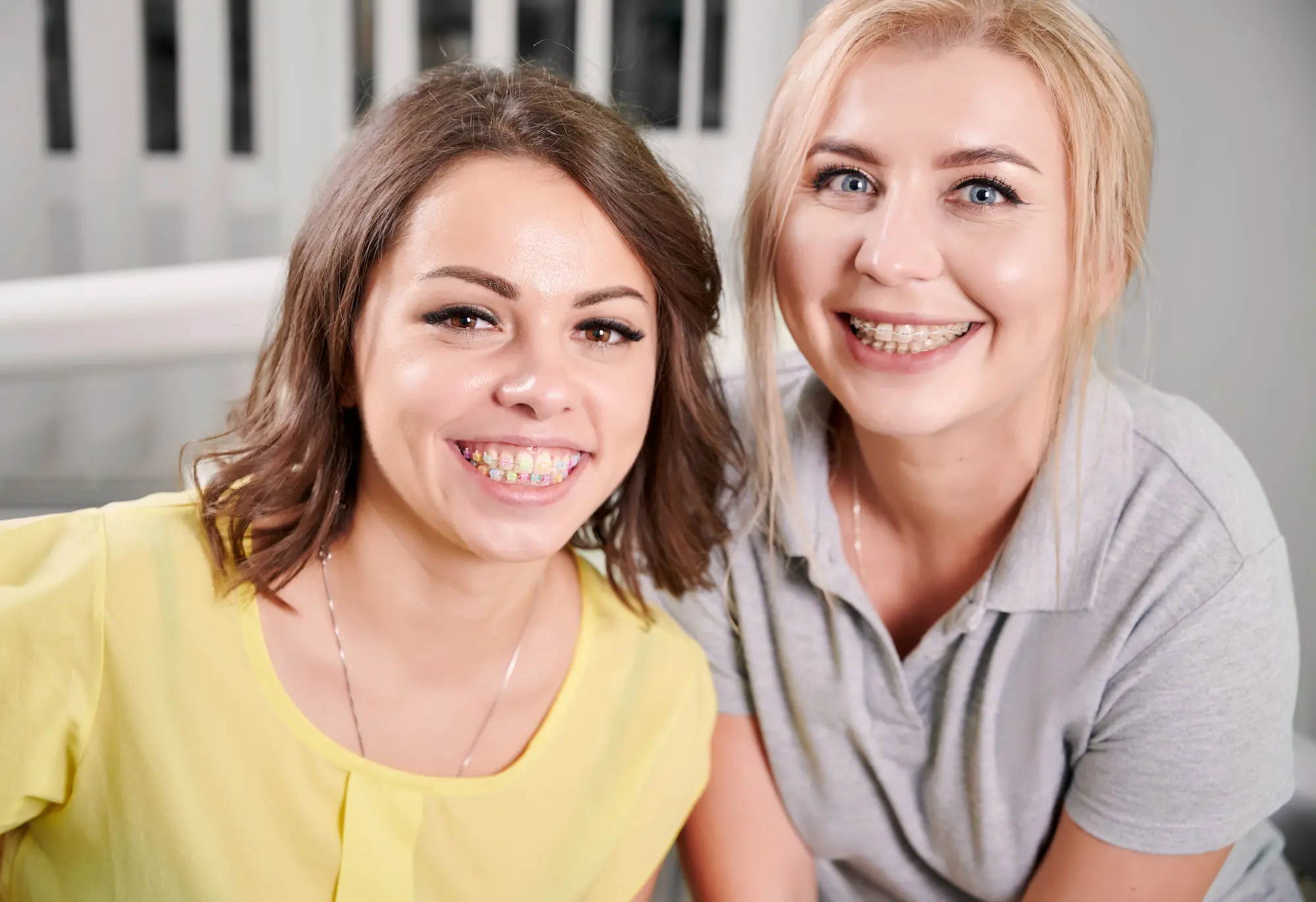 At Lenius Orthodontics in Overland Park, KS, two smiling women with affordable braces sit indoors; one wears yellow, the other gray.