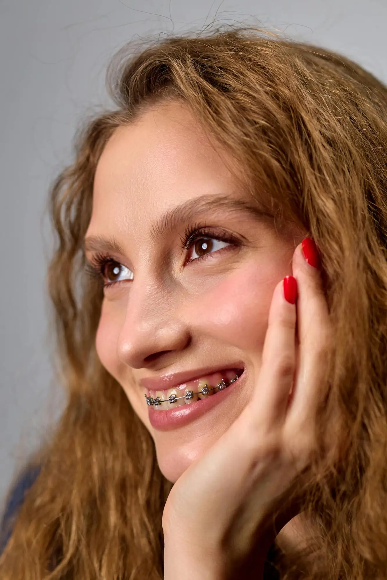 Smiling young woman with wavy hair and red nails shows metal braces for fix overbite at Lenius Orthodontics in Overland Park, KS.