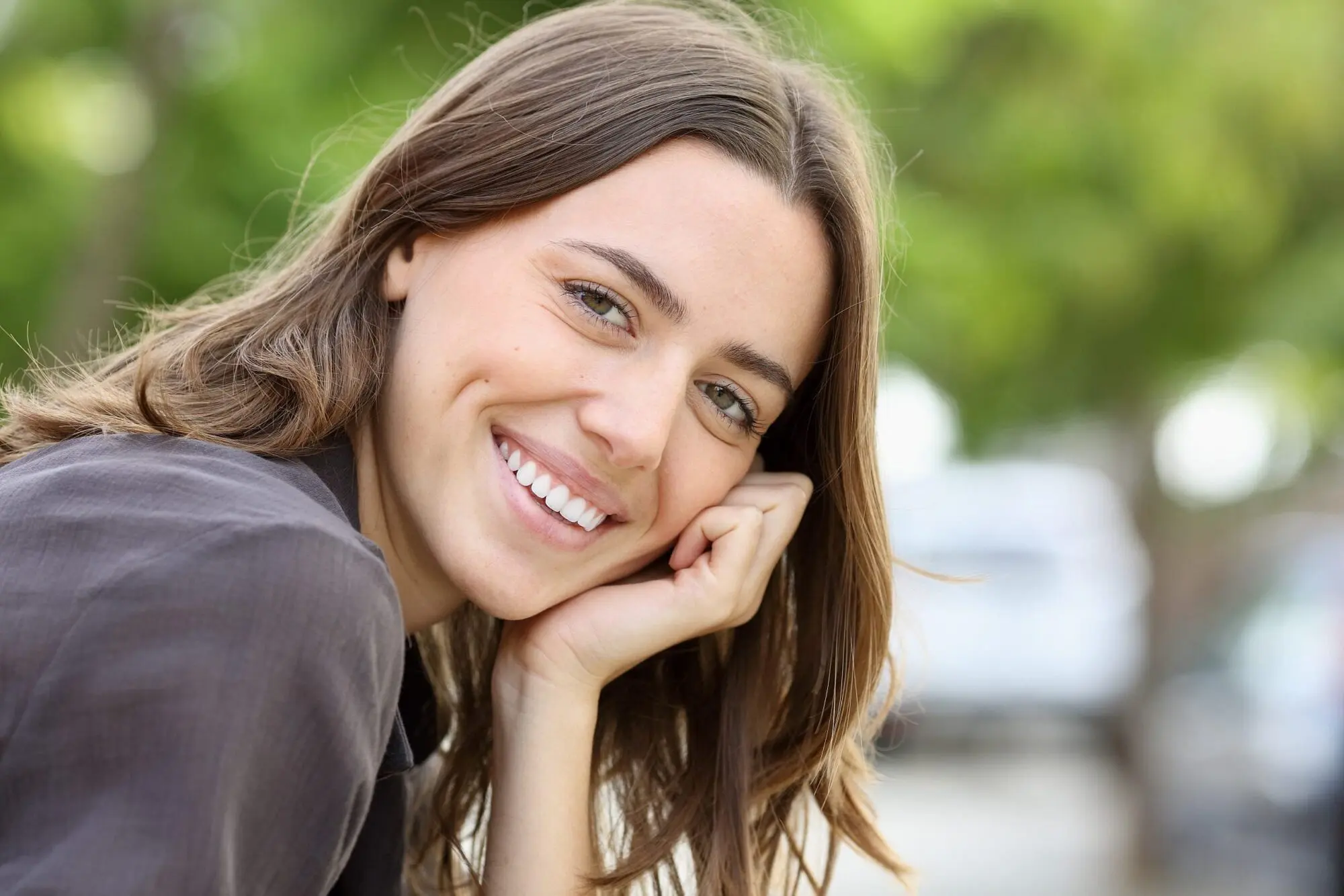 Smiling woman with long brown hair, chin resting on hand, green background blurred represent affordable braces at Lenius Orthodontics in Overland Park, KS.