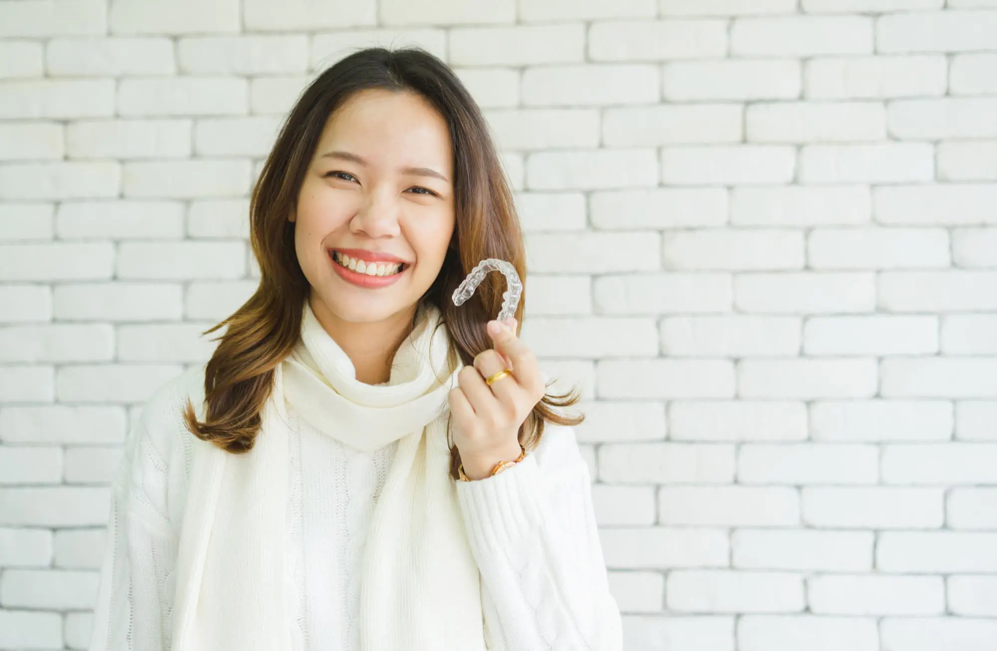 Smiling woman in a white sweater and scarf holds a clear aligner Invisalign at Lenius Orthodontics in Overland Park, KS.