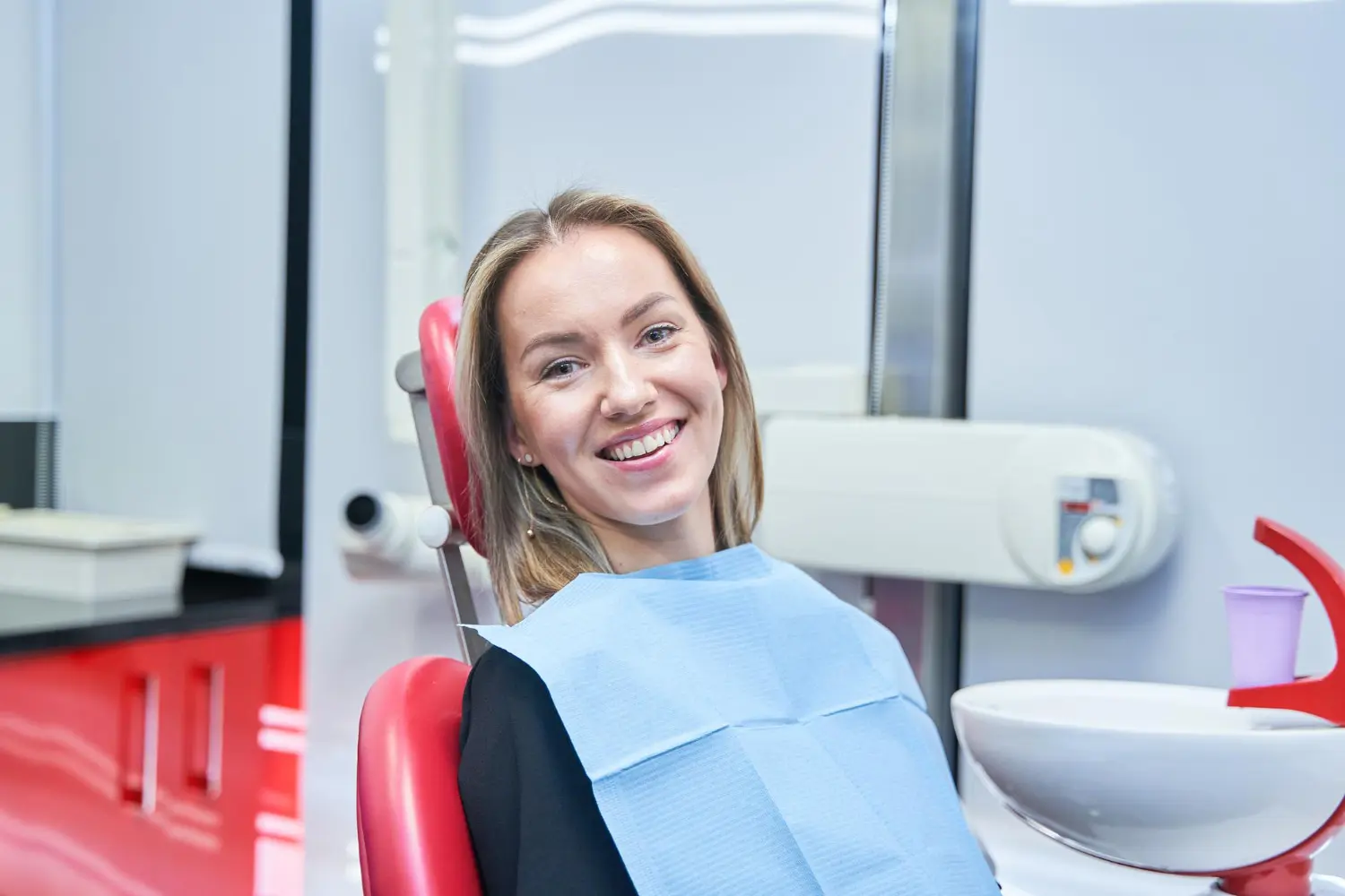 A smiling woman shows results of clear braces in a dental chair at Lenius Orthodontics in Overland Park, KS, wearing a blue bib.