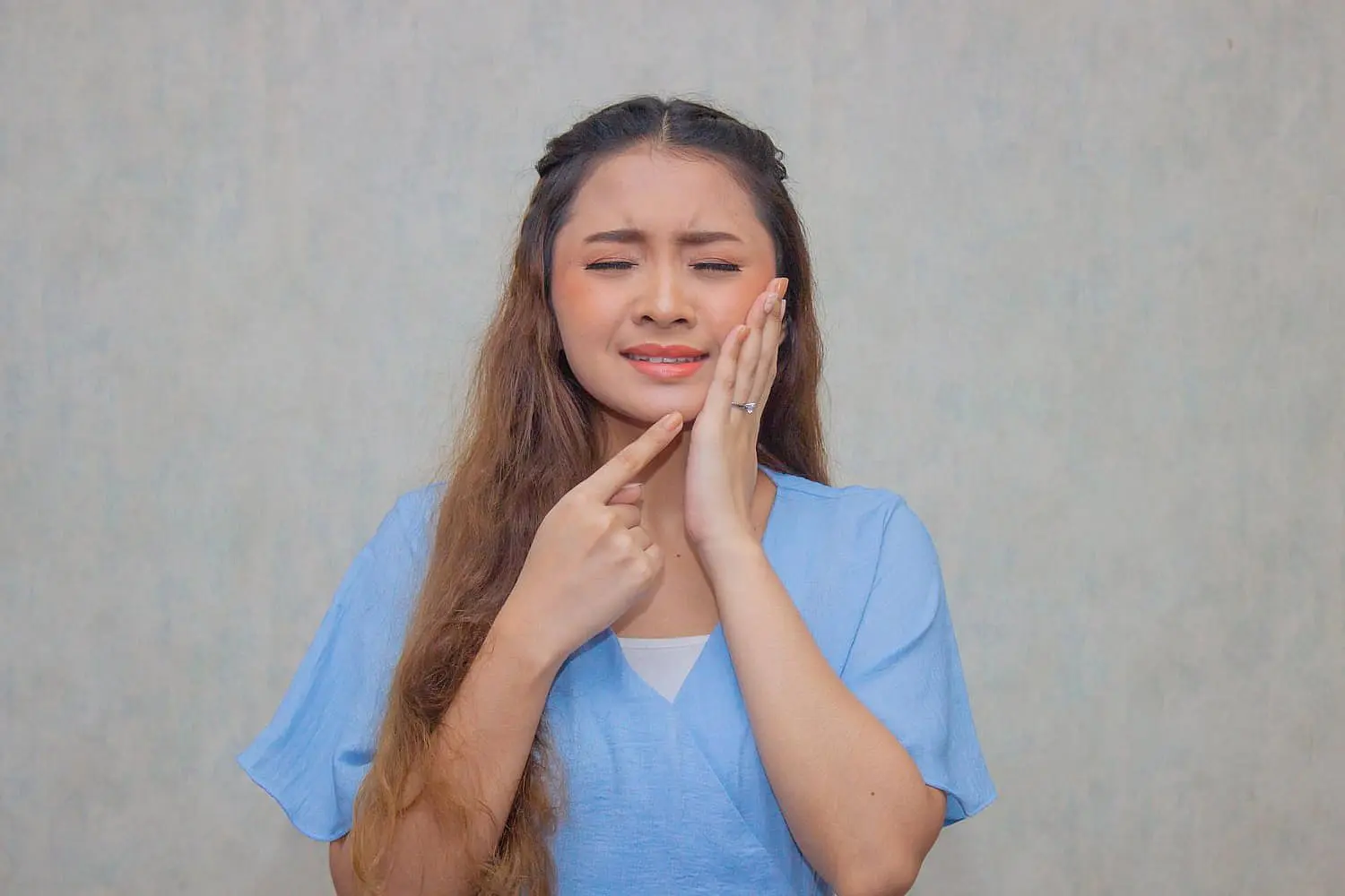 A woman in a blue shirt, appearing to have jaw pain, points to her cheek, representing genetic factors of underbite at Lenius Orthodontics in Overland Park, KS.