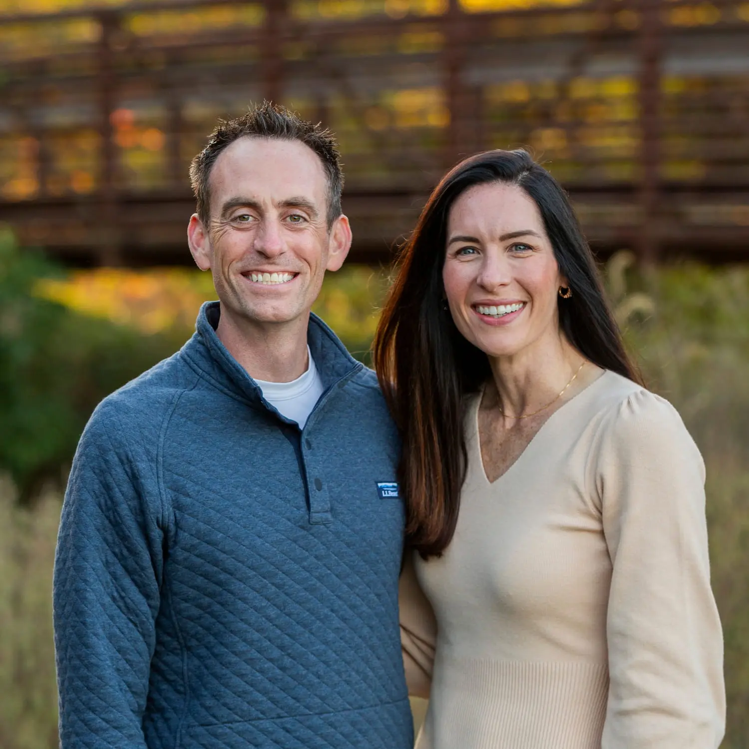 A smiling Dr. Jeffrey Lenius and his wife stand outdoors near a wooden bridge, and autumn leaves represent affordable pricing and flexible payment options for Invisalign aligners for teeth offered by Lenius Orthodontics in Overland Park, KS.