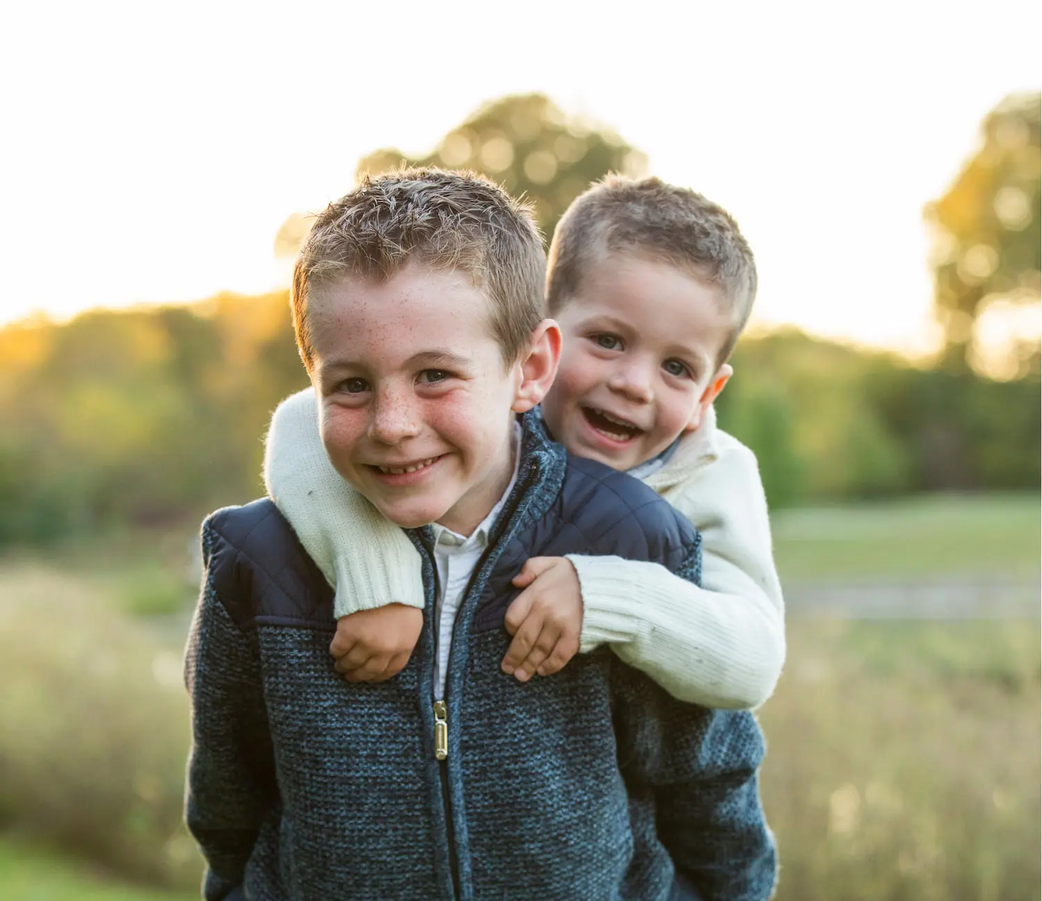 Two smiling boys outdoors, one giving the other a piggyback ride with greenery behind them, represent the best orthodontic appliances at Lenius Orthodontics in Overland Park, KS.